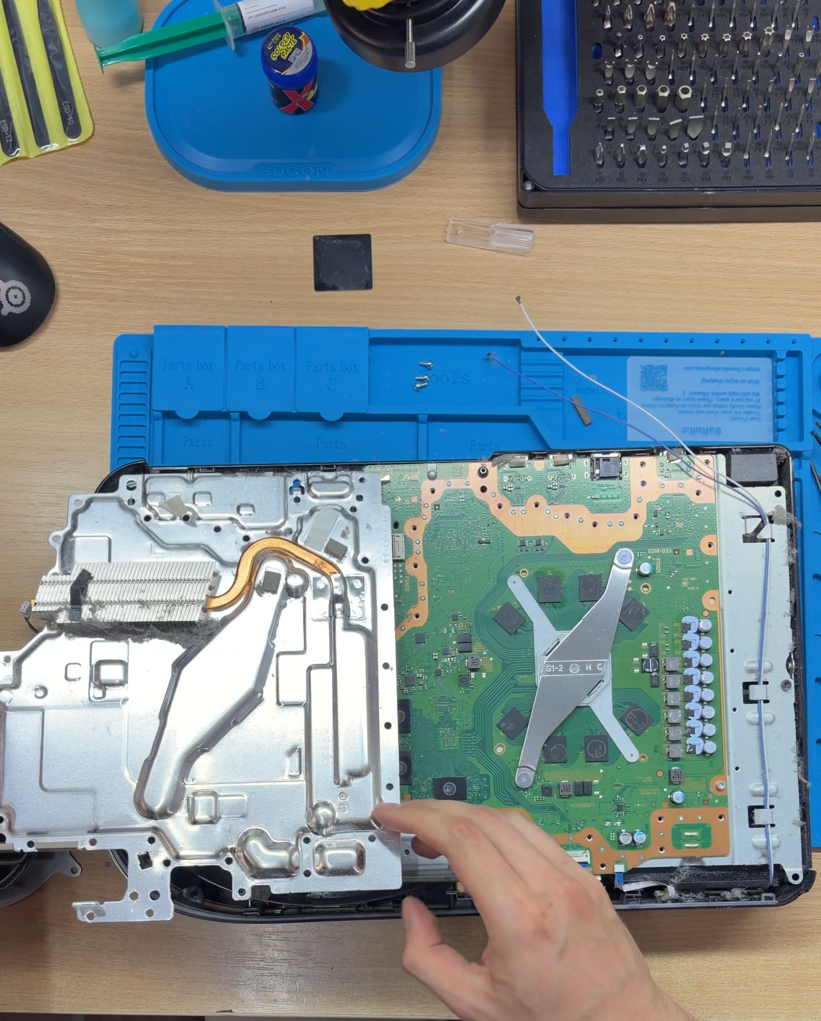 Rozłożona konsola PlayStation 5 z wyjętym układem chłodzenia; dłoń serwisanta nad odsłoniętą płytą główną